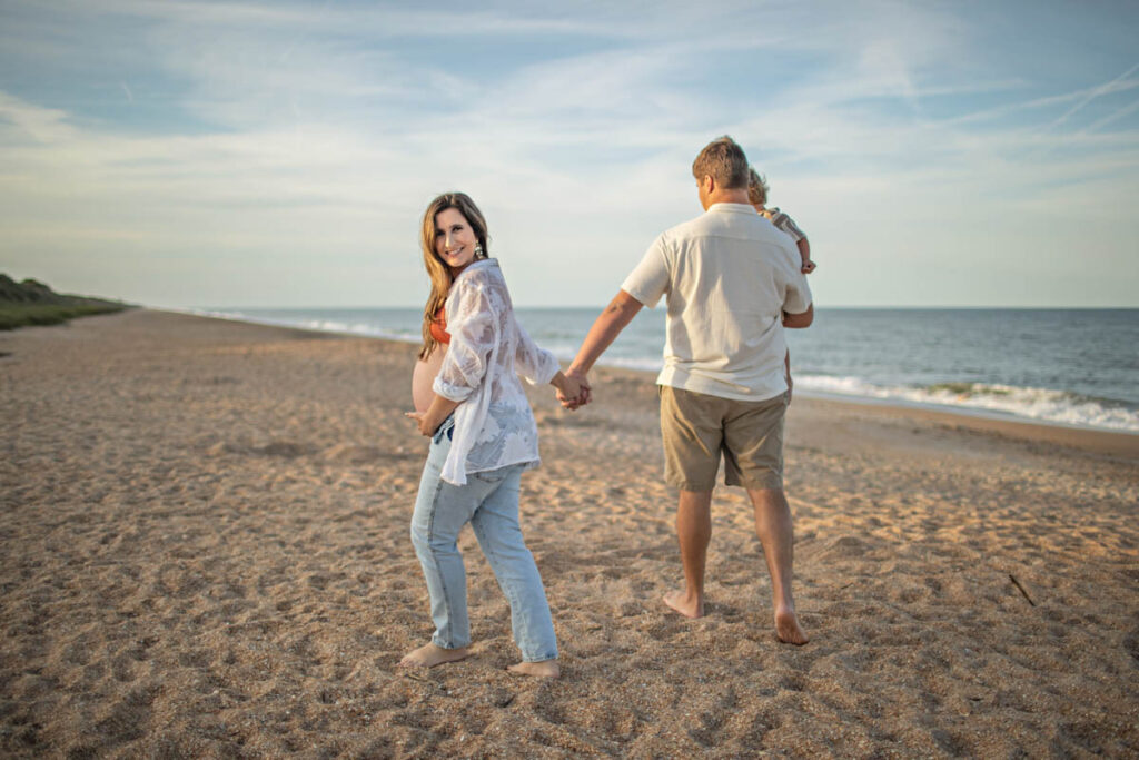 family at the beach with pregnant mother holding hands with father who is holding their toddler